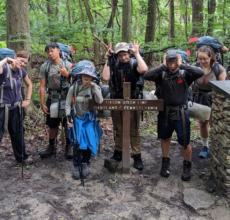 A group of seven hikers poses at a trail marker. The marker reads "MASON DIXON LINE MARYLAND / PENNSYLVANIA". The hikers are wearing backpacks and hiking gear. Some are making silly faces. The setting appears to be a wooded area. The ground is muddy.